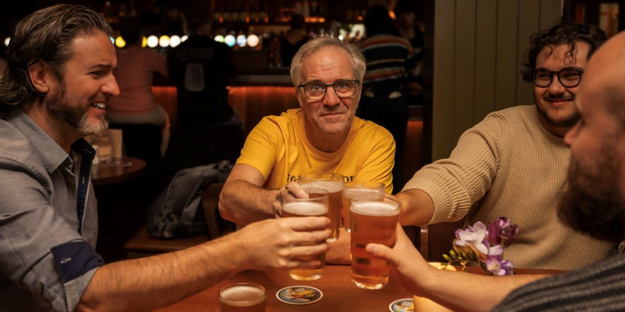 Yellow top, crafft beer,3 men, dim lighting,beard,cheers,wooden, table, lights